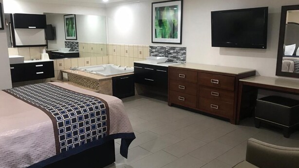 A modern hotel room with a large bed, a sitting area with a chair, a small table, a bathtub, a vanity with a mirror, and a flat-screen TV mounted on the wall.