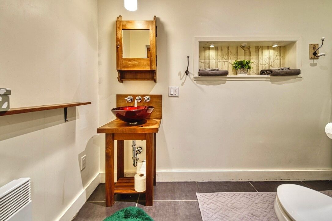 A bathroom with a wooden vanity, a red basin sink, a mirror, a window with a view of trees, and a towel rack.