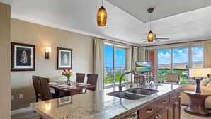 Une cuisine moderne dotée de plans de travail en granit, d’un four encastré et d’un grand îlot central avec un évier. On y trouve deux suspensions lumineuses, un ventilateur de plafond et un téléviseur posé sur un meuble. La pièce est baignée de lumière grâce à de grandes fenêtres qui offrent une vue sur l’extérieur.