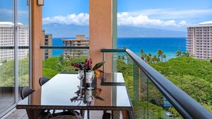 Un balcon avec une table en verre, deux chaises et un vase rempli de fleurs. Une bouteille de vin est posée sur la table. Le balcon offre une vue sur l’océan et les palmiers.
