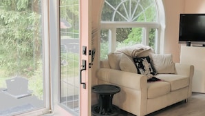 Un salon avec un canapé beige, une petite table d’appoint noire, une grande fenêtre en arc de cercle et un téléviseur à écran plat posé sur un meuble blanc.