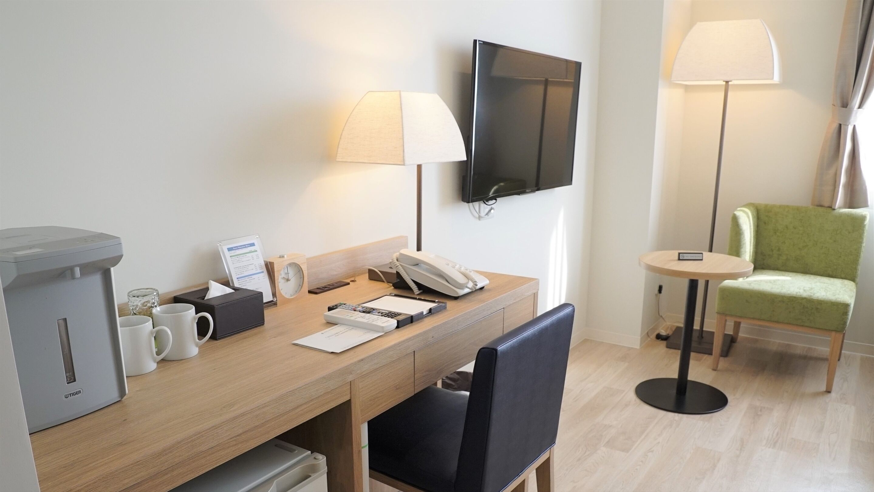 A hotel room with a wooden desk, a flat-screen TV, a telephone, a coffee machine, a green armchair, and a small round table with a lamp.