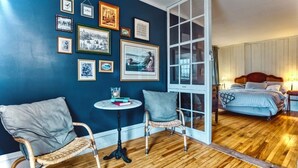 A room with a blue wall, wooden flooring, wicker chairs, a small round table, and a bed with a headboard. - Family Winter House in the ❤️ of the Eastern Townships (Ayer's Cliff)