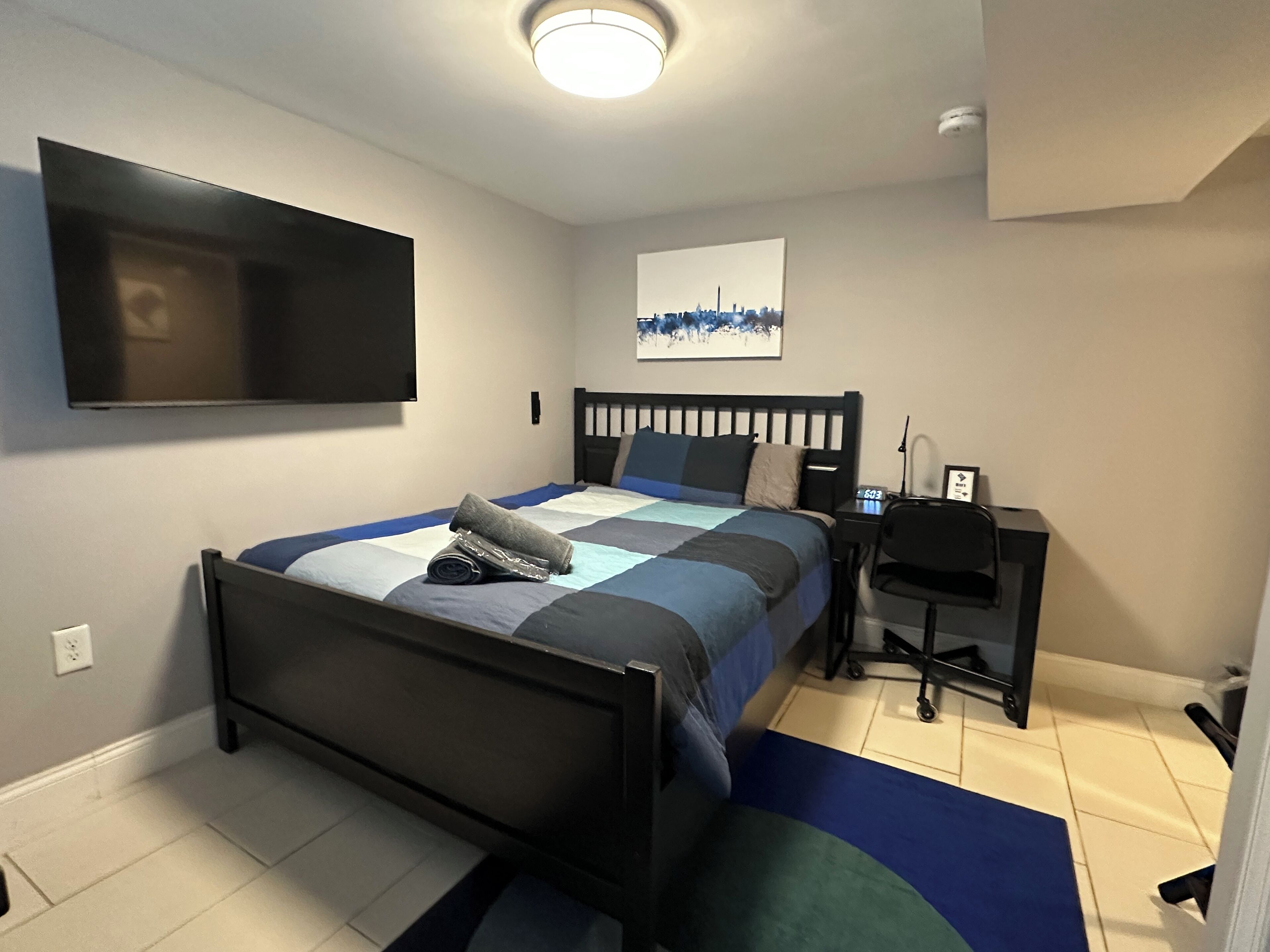 A bedroom with a bed, a desk, a chair, a TV mounted on the wall, and a painting.