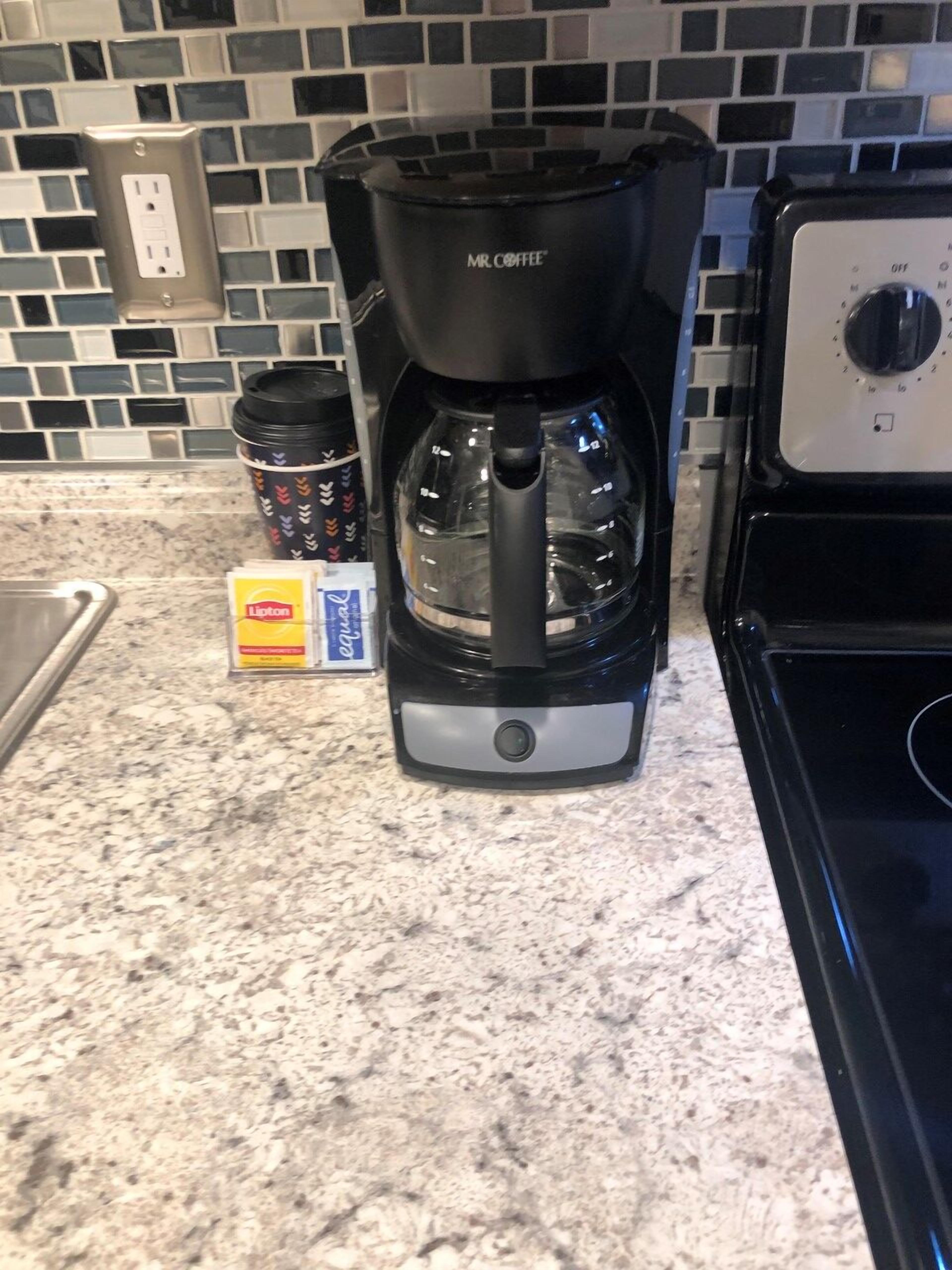 Une machine à café de la marque Mr. Coffee est posée sur un plan de travail de cuisine, avec un mur recouvert de carreaux et une cuisinière.