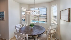 Un comedor con una mesa redonda de madera, sillas blancas, una lámpara colgante y ventanas amplias con persianas.