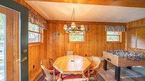 Habitación con piso de madera, una mesa y sillas de madera, una mesa de fútbol y una ventana con cortinas a cuadros.
