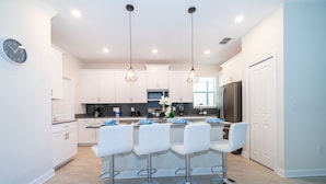 Une cuisine moderne avec des meubles blancs, un îlot central doté de tabourets, une horloge et des luminaires suspendus.
