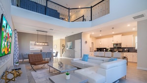 Un vaste espace de vie comprenant un canapé blanc, un fauteuil marron et une table basse en verre. Un escalier permet d’accéder à l’étage supérieur.