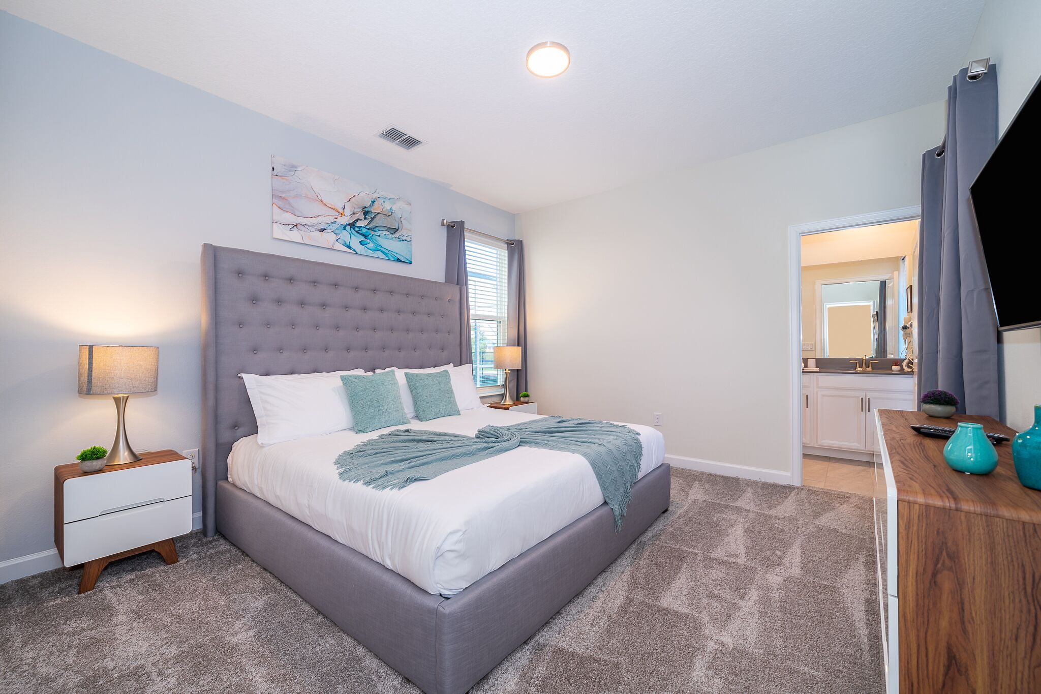 A bedroom with a large bed, a nightstand, a lamp, a painting on the wall, and a view into the bathroom.