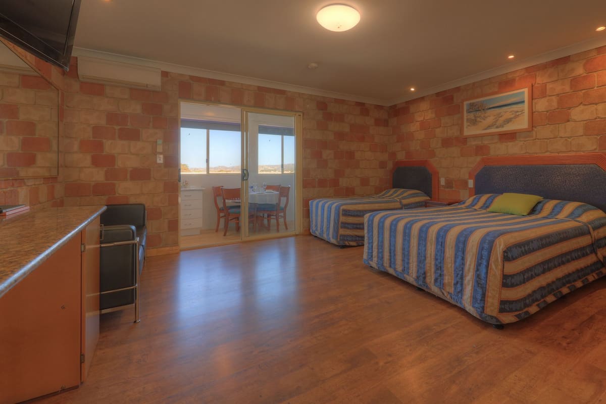 A room with two beds, a dining table, chairs, a kitchen area, and a sliding glass door leading to an outdoor area.