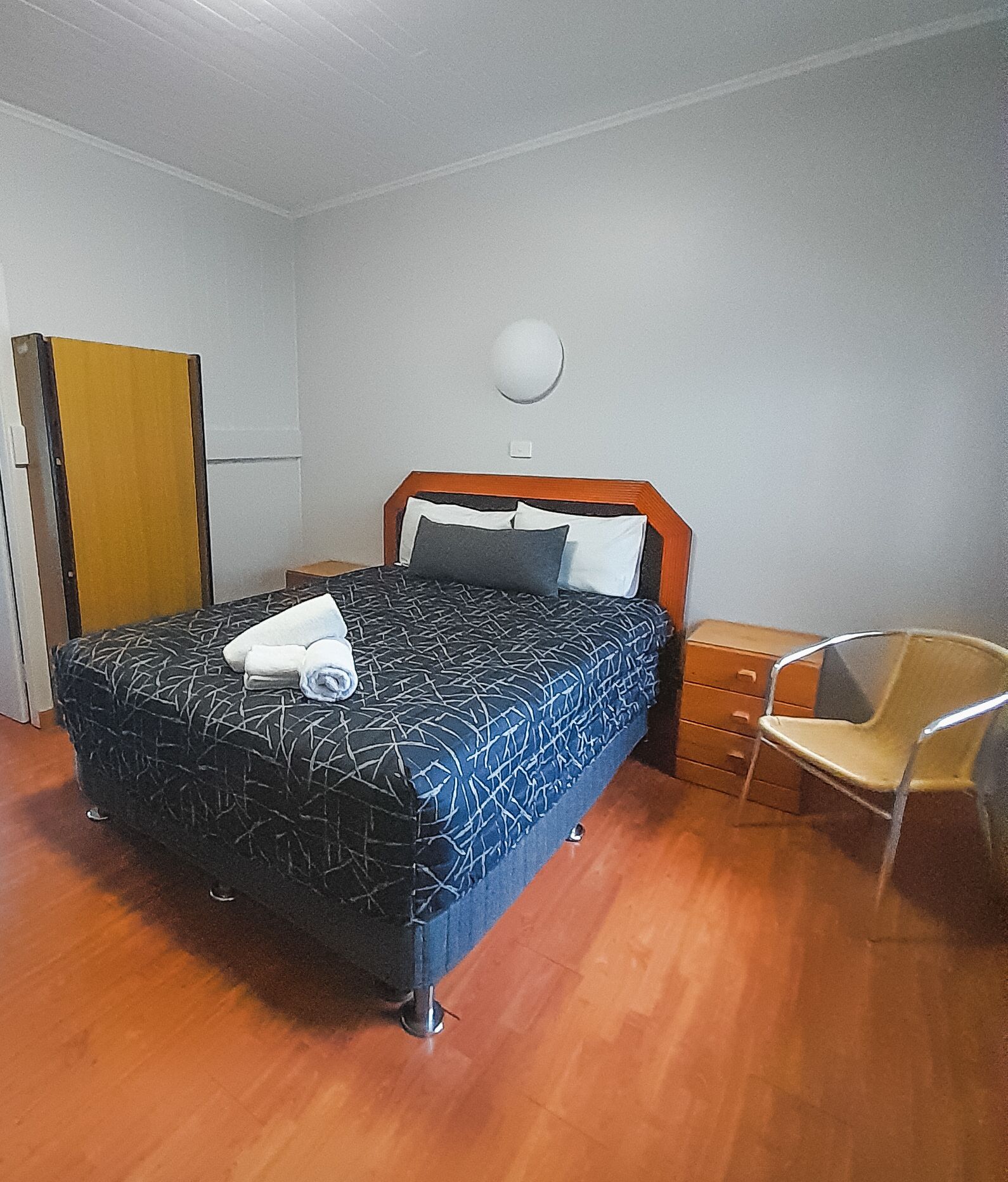 Ein ordentlich bezogenes Bett mit einem gemusterten Bettüberwurf und Kissen, ein Nachttisch aus Holz mit Schubladen, ein Stuhl und eine runde Wandlampe.