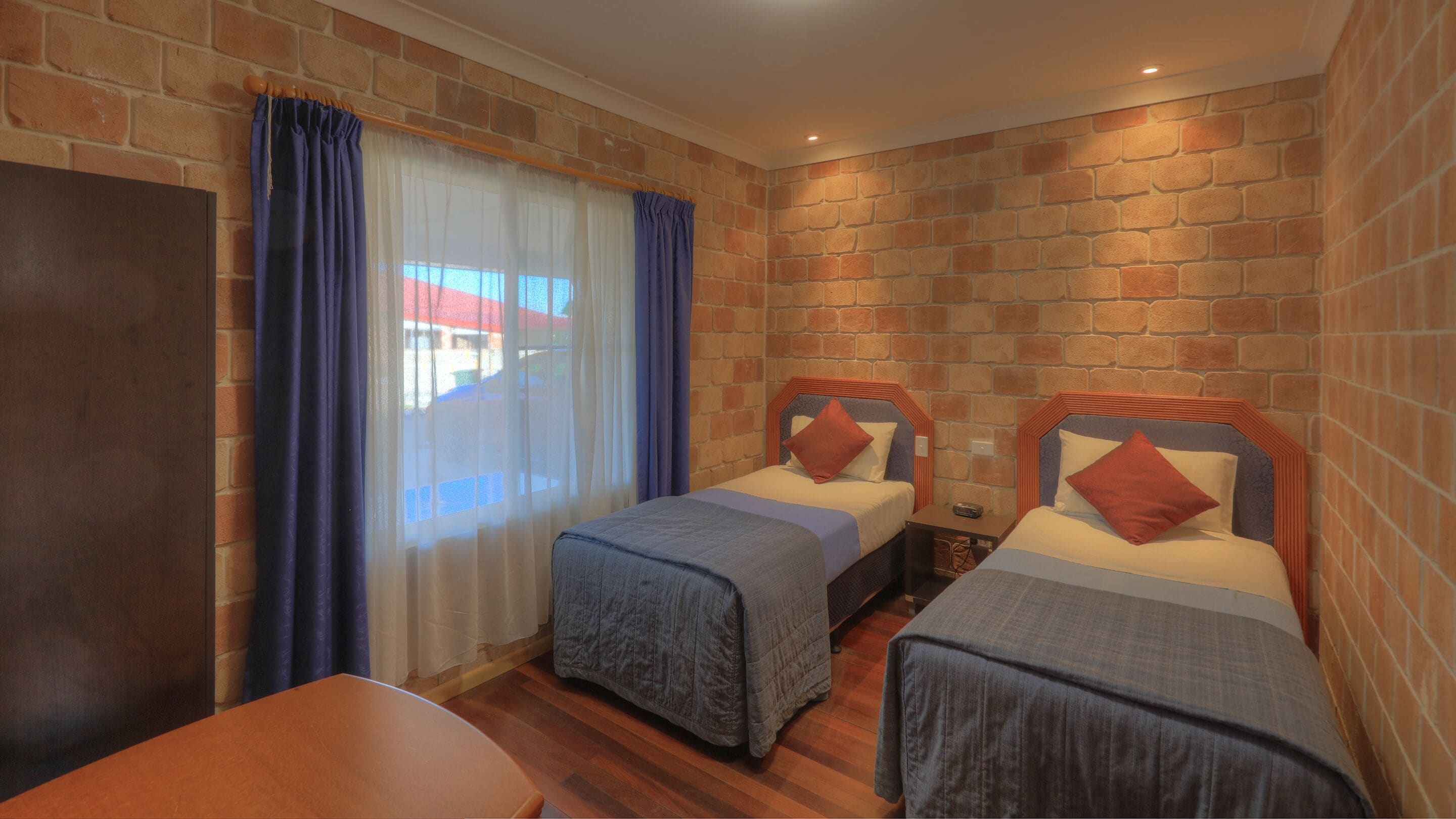 A room with two single beds, a wooden table, and a brick wall.