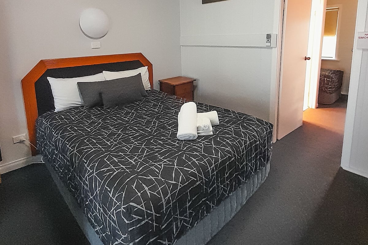 A bedroom with a bed, a bedside table, a wall-mounted lamp, and a door leading to another room.