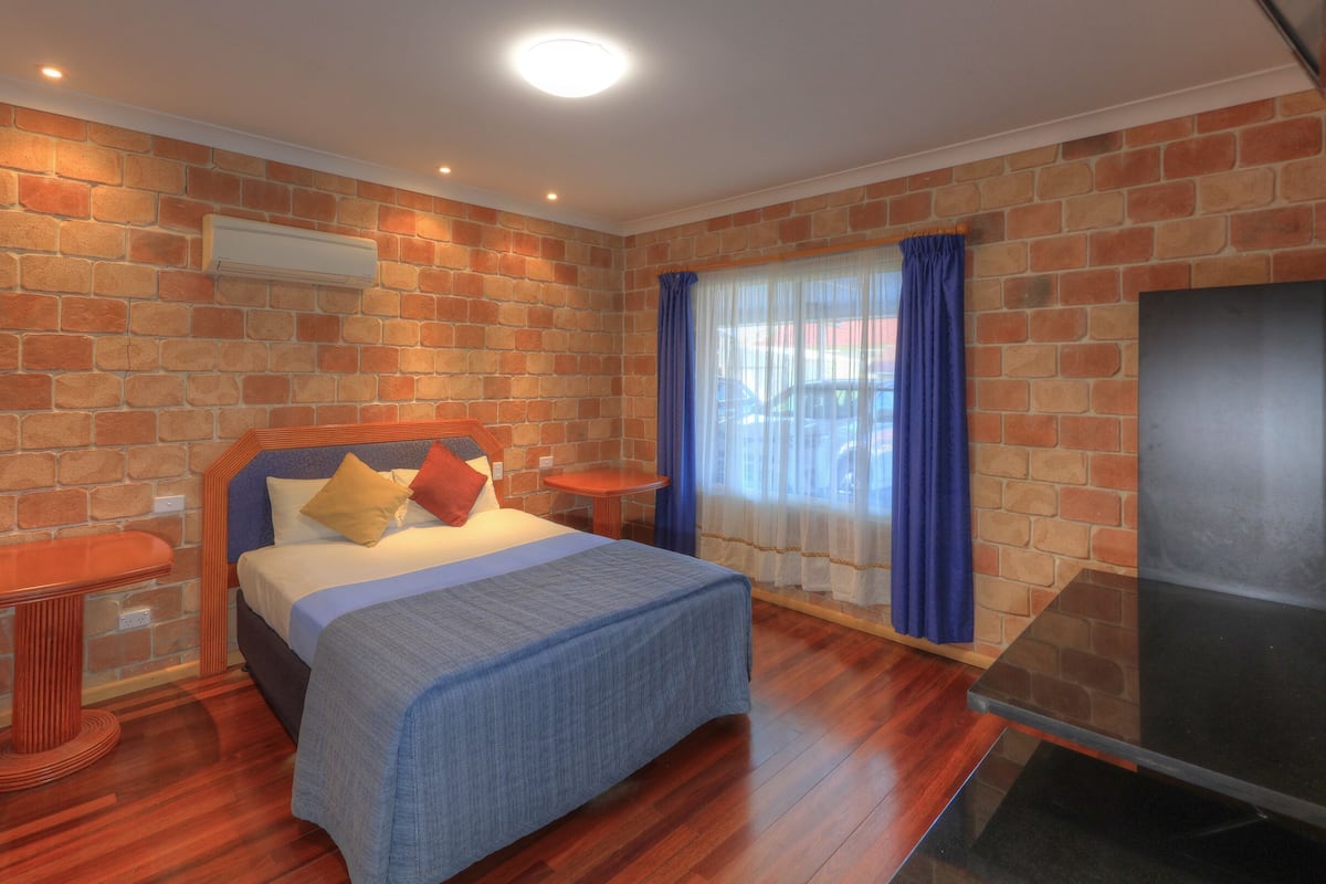 A bedroom with a brick wall, a bed with pillows, a small table, and a TV.