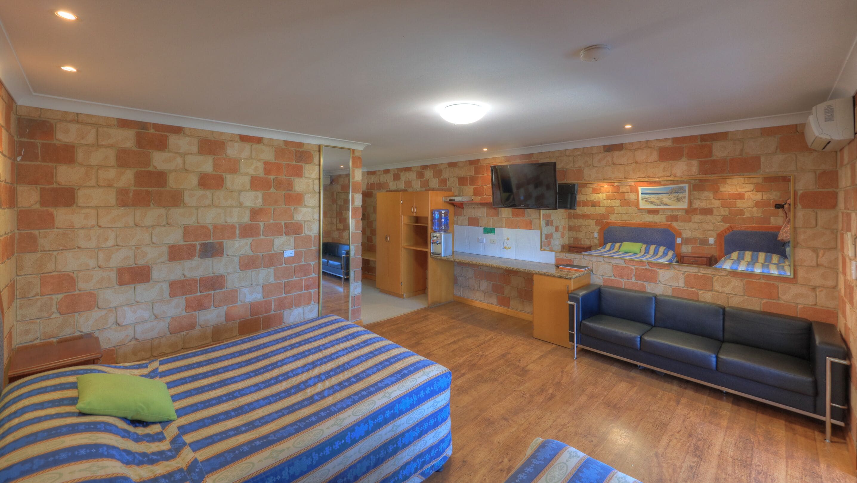 Una habitación compacta con cama, sofá, kitchenette pequeño y televisor montado en la pared.