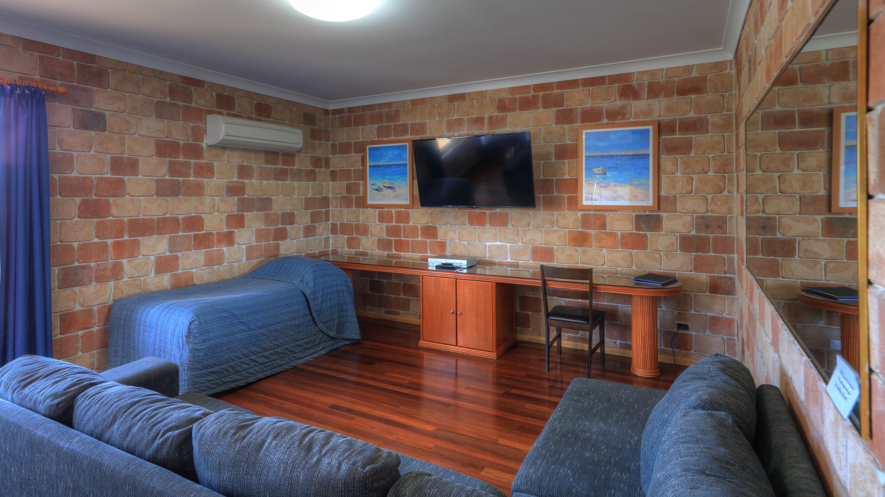 A room with a brick wall, a flat-screen TV mounted above a desk, a bed with a blue cover, and a sofa.