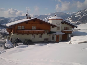 A two-story chalet with a snow-covered roof, a balcony, and a mountain backdrop.