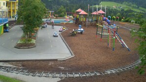 A playground with various play structures, a small parking area, and a building in the background.