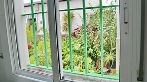 A window with a green metal grid, through which a garden with various plants and a white building is visible.