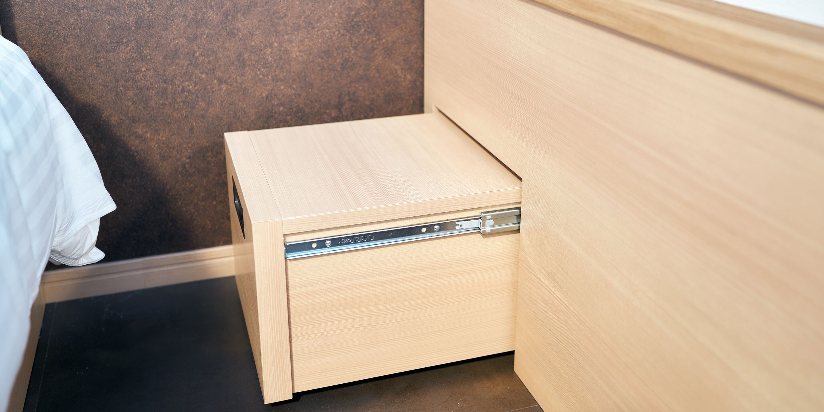 Une petite table de chevet en bois, dotée d’un tiroir, placée à côté d’un lit dans une pièce dont un mur présente une texture sombre.