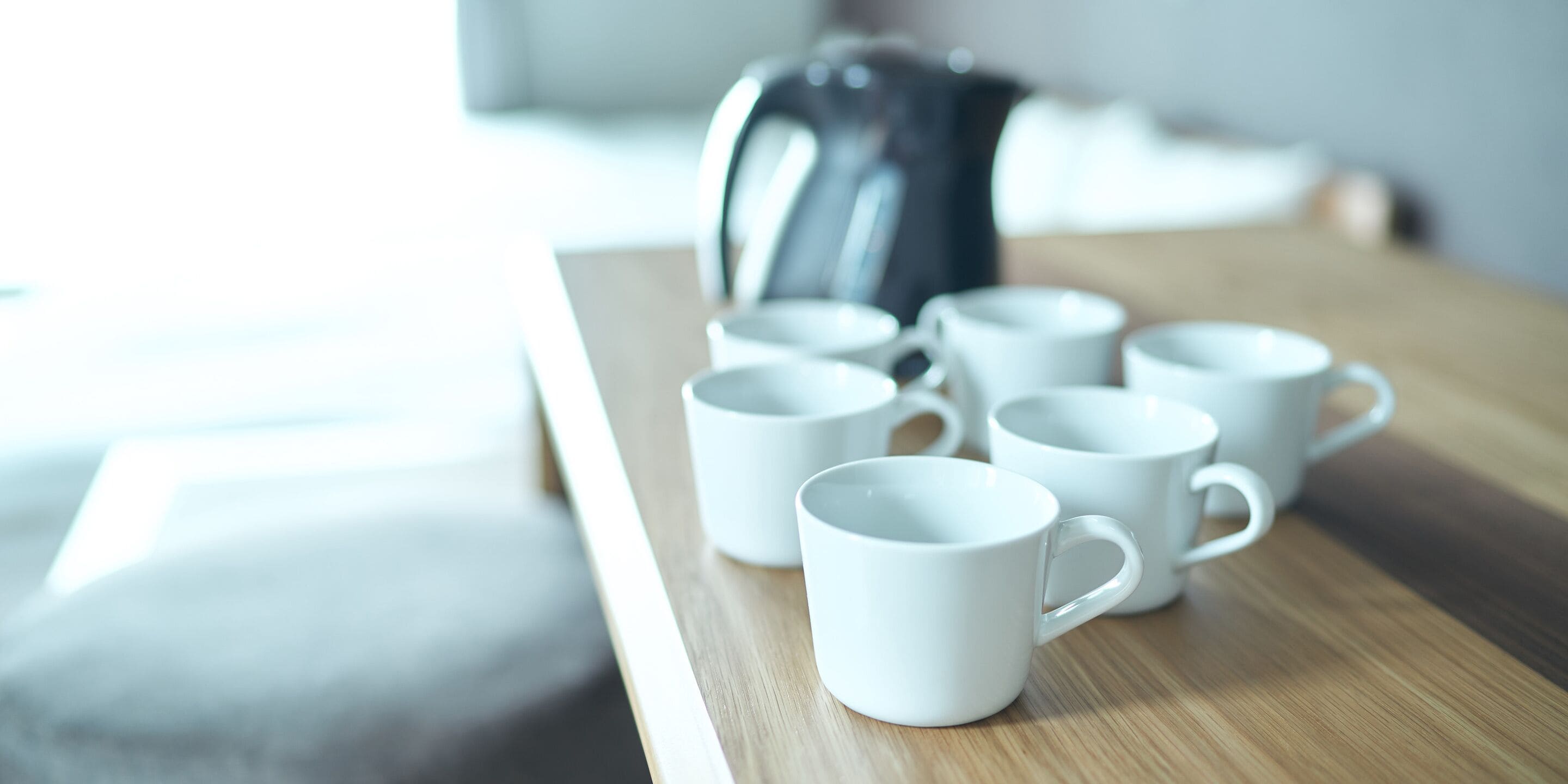 Una mesa de madera con seis tazas blancas de cerámica y una tetera eléctrica negra.