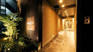 L’entrée d’un hôtel, avec une enseigne indiquant « Rakuten Stay », une façade de bâtiment moderne et un chemin bien éclairé menant à l’intérieur.