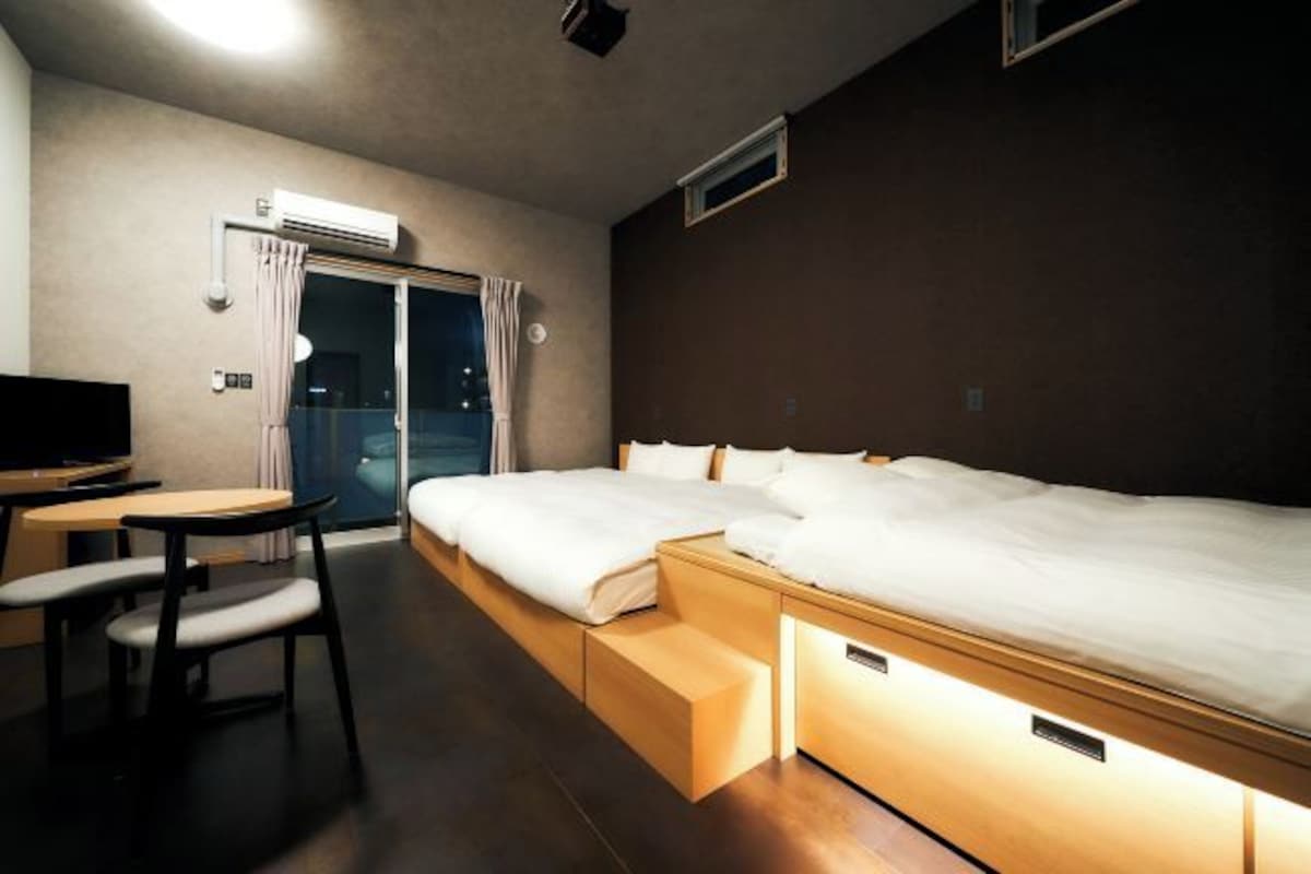 A hotel room with a bed, a desk, and a chair. There is a window with curtains, an air conditioning unit, and a small table.
