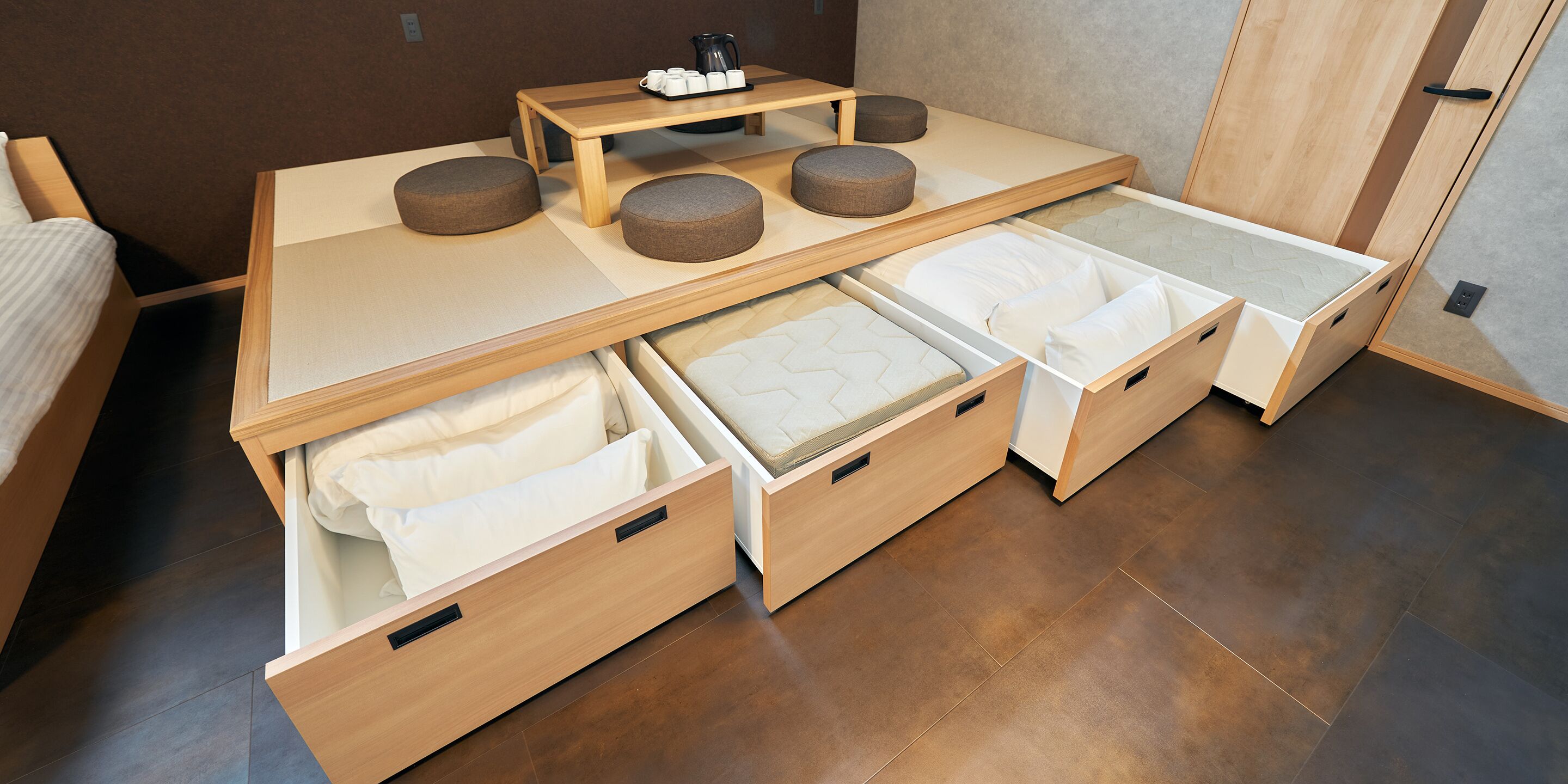 Una habitación compacta y multifuncional con cama, cajones para guardar objetos, una mesita con tazas y hervidor, y un rincón de descanso con cojines.