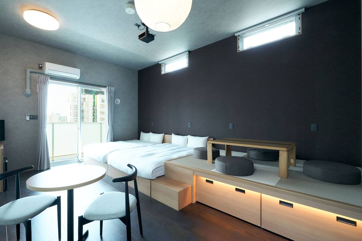 A modern hotel room with a bed, bedside tables, a TV, and a seating area with chairs and a table.