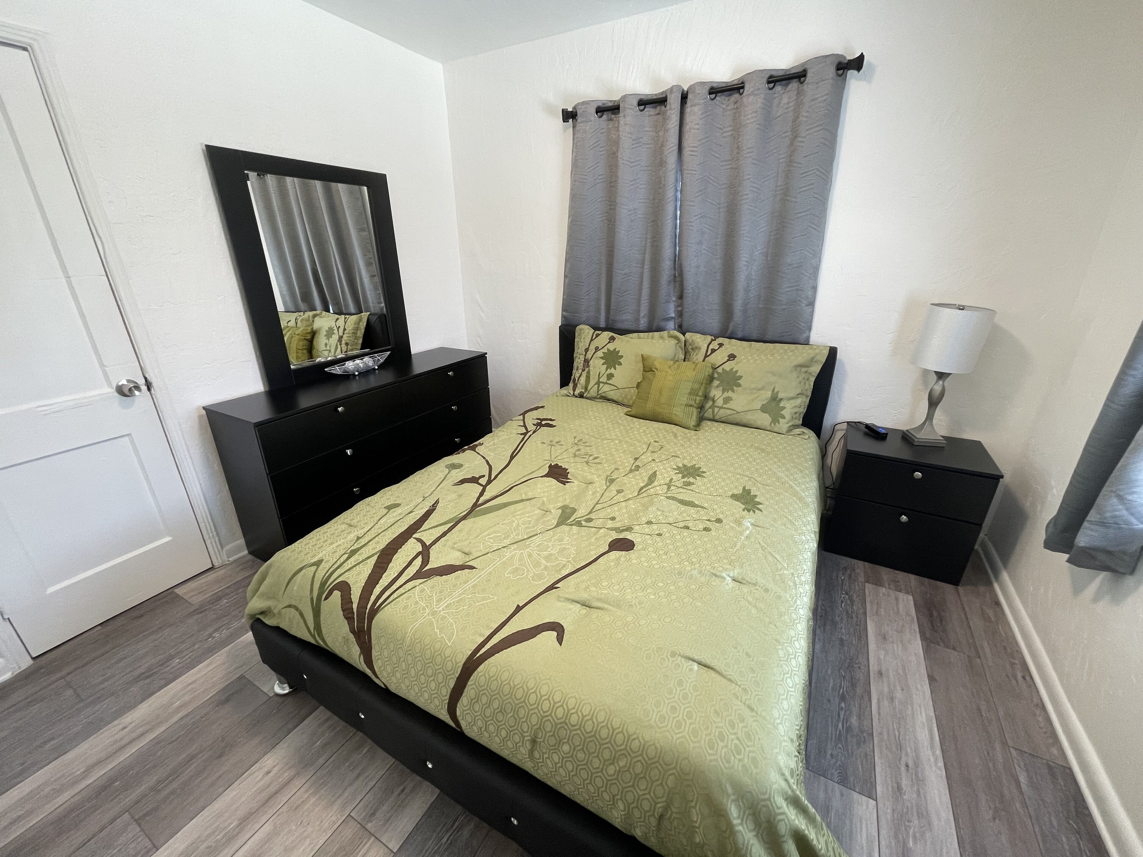 Une chambre à coucher comprenant un lit, une commode, une table de chevet, une lampe, un miroir et une porte.