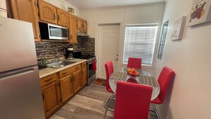 A compact kitchen with wooden cabinets, a stainless steel refrigerator, a microwave, and a coffee maker. There is a dining area with a glass table and red chairs.