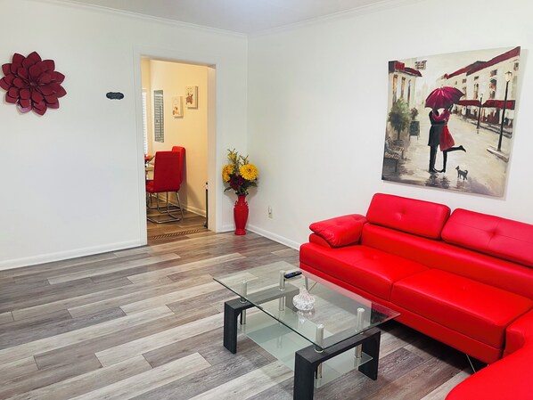A modern living room with a red sofa, a glass coffee table, and a painting of a street scene.