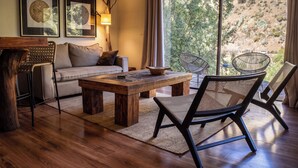 A modern living room with a wooden coffee table, a sofa, and two chairs. There are two framed artworks on the wall, a lamp, and a view of a natural landscape through the window. - House on the way to Farellones (Santiago)