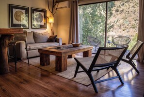 Una sala de estar moderna con una mesa de centro de madera, un sofá y dos sillones. Hay dos obras de arte enmarcadas en la pared, una lámpara y vistas de un paisaje natural a través de la ventana.