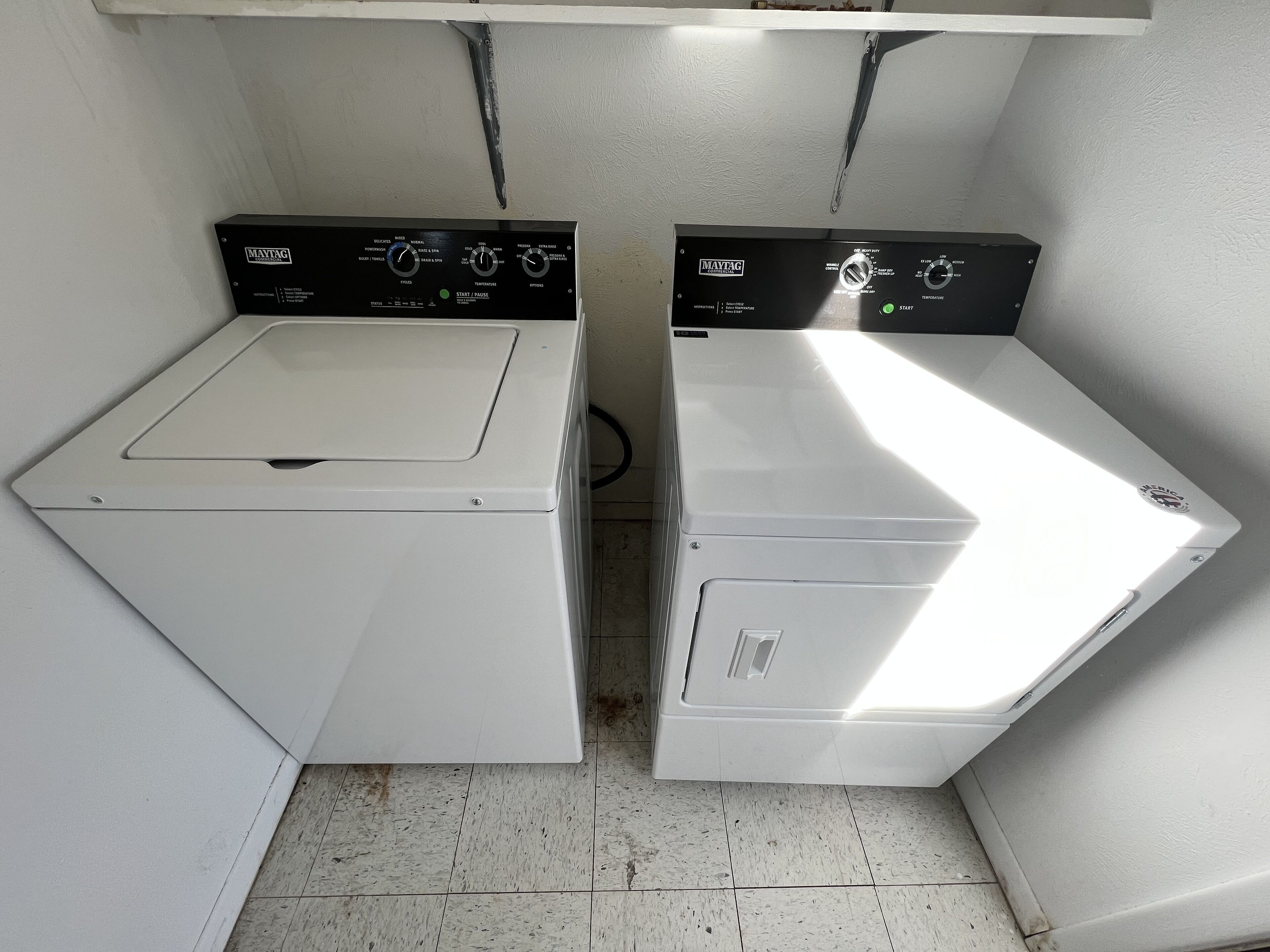 Deux machines à laver à chargement frontal, de couleur blanche, sont placées côte à côte dans une buanderie dont le sol est carrelé.