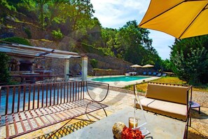 Un espace aménagé au bord de la piscine, avec un barbecue en pierre, des sièges d’extérieur et une vue sur une colline naturelle.