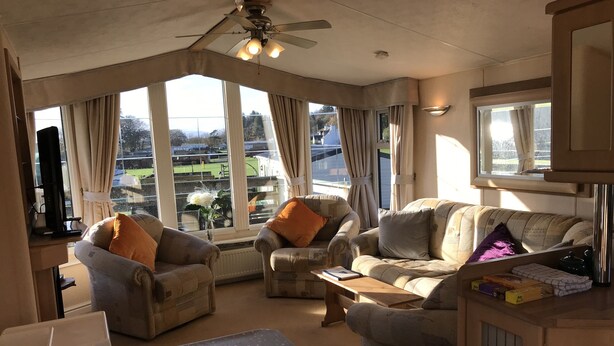 Ein gemütliches Wohnzimmer mit zwei Sesseln, einem Sofa, einem Couchtisch und einem Fernseher. Große Fenster bieten Blick ins Freie.