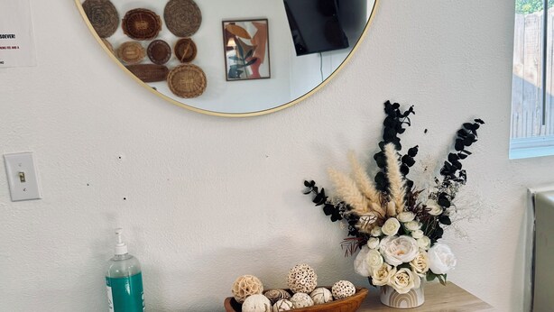 Un bureau en bois avec un miroir, un bol rempli d’objets décoratifs, une bouteille de gel hydroalcoolique et un vase de fleurs.
