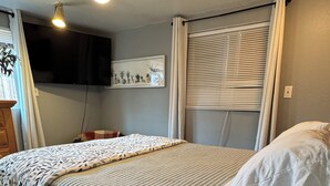 Une chambre à coucher avec un lit, un ventilateur de plafond, un téléviseur fixé au mur et une fenêtre avec des stores.
