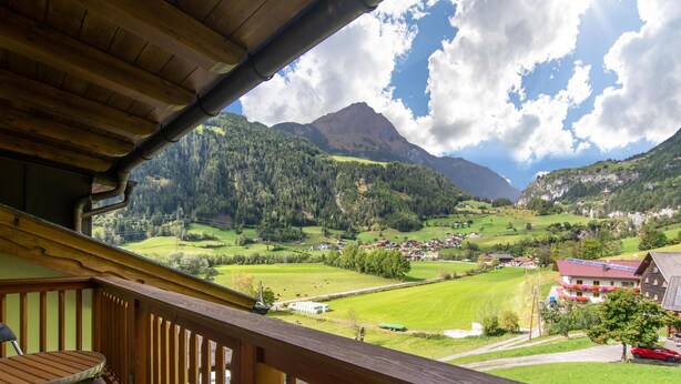 Un balcon en bois, agrémenté d’une table et de chaises, qui offre une vue sur une vallée verdoyante, avec des maisons et des montagnes.