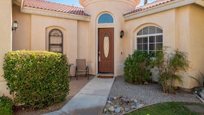 A building with a brown door, a round window, and a small patio area with a chair and table. - Private home,2 King Beds, near Empire Polo, Tennis, Pools, Coachella, Horse Pk, (Indio)