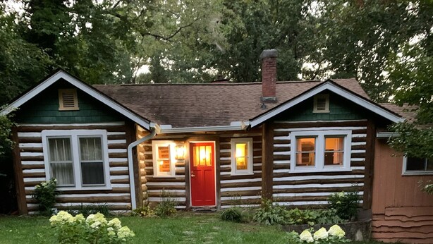 Eine Blockhütte mit roter Tür, Steintreppe zum Eingang und einem gepflegten Garten mit Hortensien.