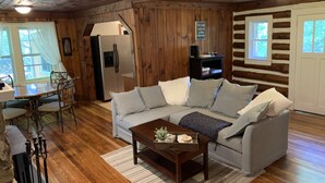 Ein gemütliches Blockhaus-Interieur mit Holzboden, einer Couch, einem Couchtisch, einem Esstisch mit Stühlen und einer Küchenzeile mit Kühlschrank.