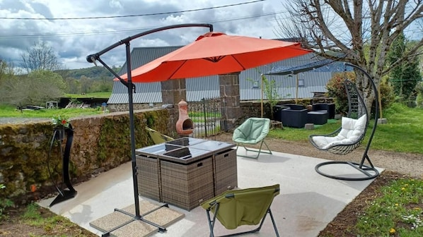 En udendørs terrasse med et fletbord, en hængestol, en gyngestol og en rød parasol, der giver skygge.
