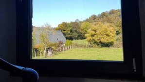A window with a view of a grassy field and trees, a stone building, and a clear blue sky. - House in Carladez with private sauna (Brommat)