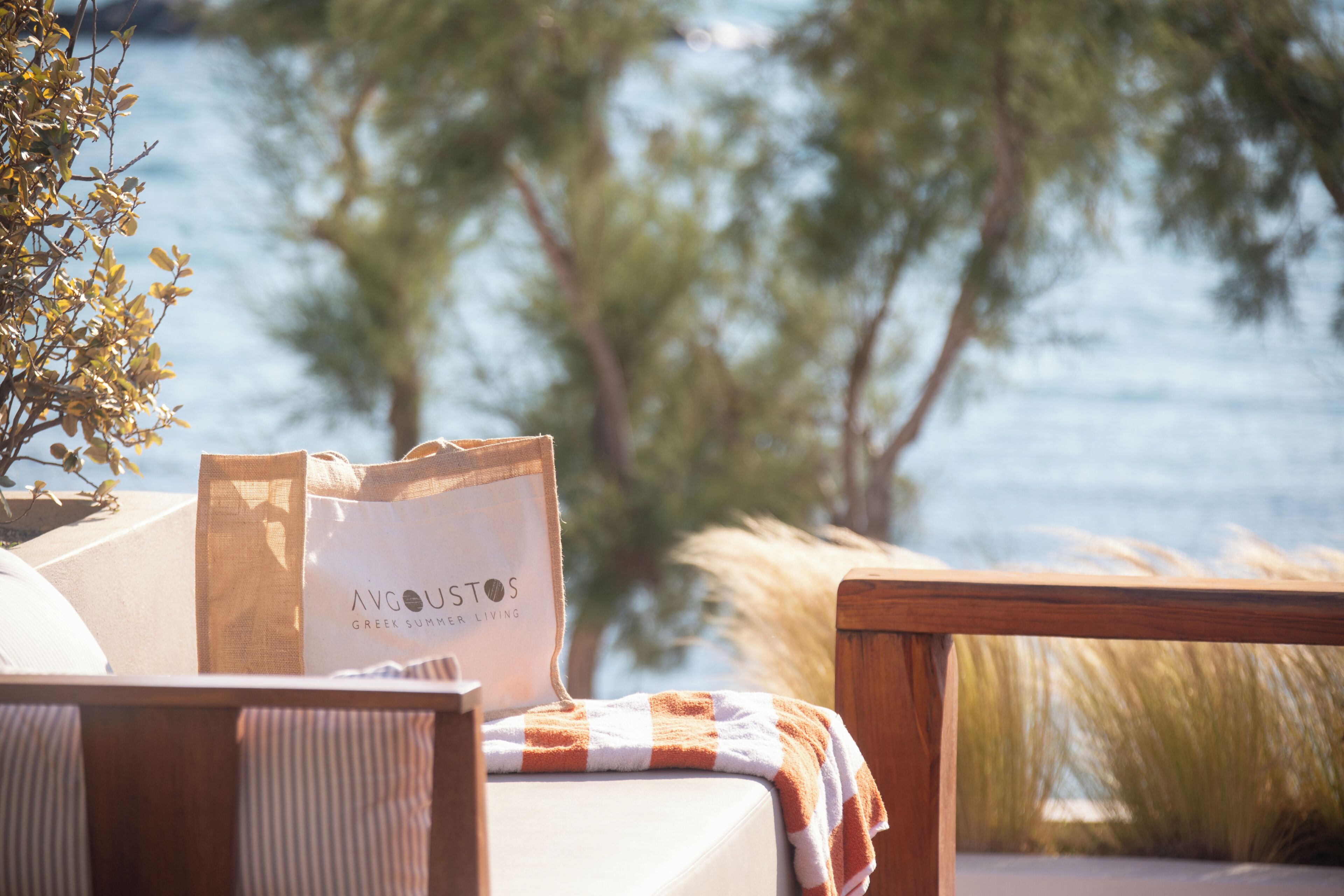 Um cenário à beira-mar com uma cama, uma bolsa com a etiqueta 'AVGOUSTOS GREEK SUMMER LIVING' e vista para o mar e árvores.