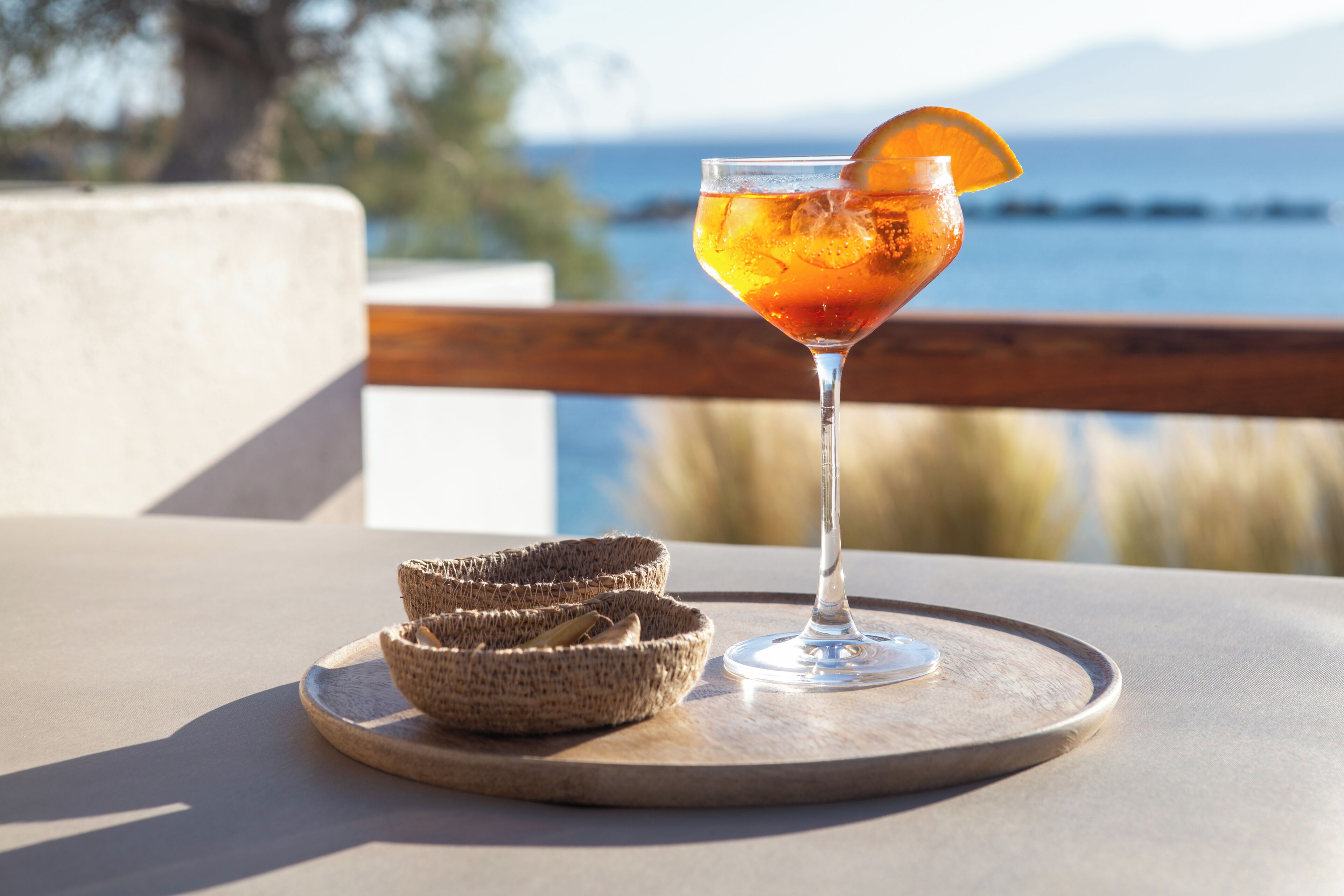 Ein Glas bernsteinfarbenes Getränk mit Orangenscheibe auf einem Holztablett, begleitet von zwei gewebten Körben, steht auf einem Tisch mit Blick auf Meer und Berge.