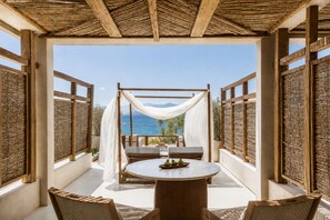 An outdoor seating area with a table and chairs, a view of the sea, and a covered structure with a woven roof. - Avgoustos Suites - Adults only (Naxos)