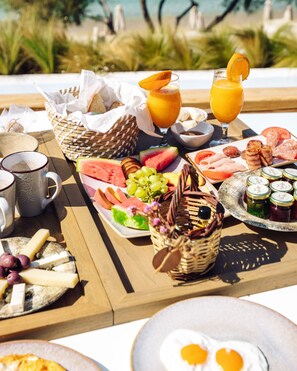 A breakfast tray with various foods and drinks, including eggs, toast, fruits, and a glass of orange juice, set on a wooden table with a view of the beach in the background. - Avgoustos Suites - Adults only (Naxos)